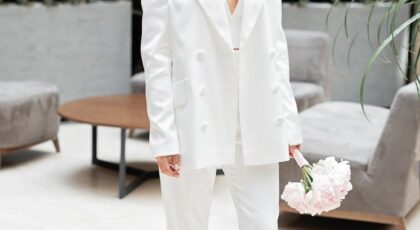 Chic and modern bride in a white suit holding a bouquet indoors. Stylish elegance.