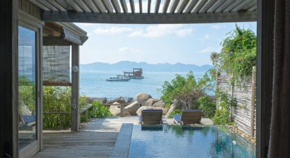 Elegant wooden deck with infinity pool overlooking serene seaside view with mountains and boat.