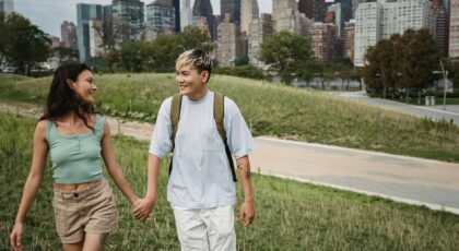 Positive diverse couple in casual apparel holding hands while walking on grass lawn near contemporary buildings and looking at each other