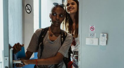 Two young women with backpacks enter a hostel room, ready for adventure.