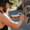 A young man adjusts a barbell outdoors while wearing headphones, focusing on fitness.