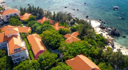 Aerial shot of red-roofed villas by a clear blue sea, surrounded by lush greenery.