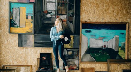 A woman holding a vinyl record stands amidst eclectic artwork and retro decor.