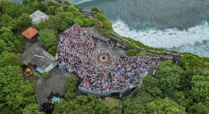 An aerial view of a crowded amphitheater at Uluwatu Temple in Bali, surrounded by lush greenery and ocean views.